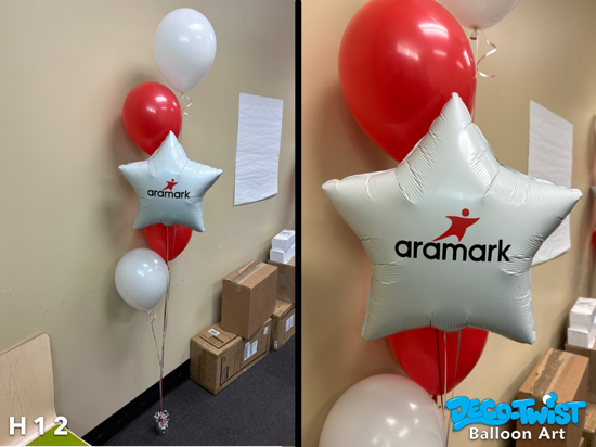This image shows a helium balloon bouquet placed indoors against a beige wall. The arrangement includes red and white latex balloons along with a white star-shaped foil balloon featuring the Aramark logo in custom vinyl. The balloons are tied together with curling ribbons and anchored to a small weight on the floor.