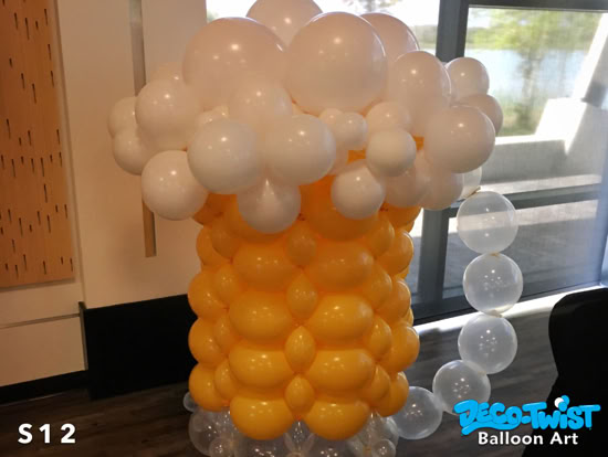A balloon sculpture shaped like a giant mug of beer is shown, with yellow balloons forming the glass and clusters of white balloons on top to look like foam.