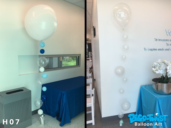 This image shows two elegant helium balloon bouquets with a cascading design: On the left, a large white balloon is at the top, with a vertical strand of smaller balloons in shades of blue, white, and clear, creating a floating bubble effect. They are anchored with a silver balloon weight. On the right, a large clear balloon tops the bouquet, followed by a strand of smaller clear balloons in descending sizes, resembling floating bubbles. This one is also anchored with a small weight at the bottom. Both bouquets have a sleek, modern look, ideal for formal or upscale events.