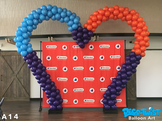 A large heart-shaped balloon arch stands in front of a red backdrop with the “Devoted Health Plans” logo. The left side of the arch is made of blue and purple balloons, while the right side is orange and purple, meeting at the top to form a heart shape. It creates a bold and colorful display for the event.