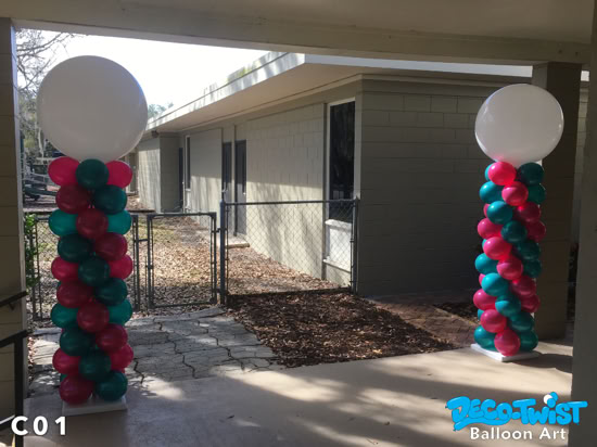 Two tall balloon columns stand side by side outdoors, each topped with a large white round balloon. The columns are spiraled with alternating teal and magenta balloons, creating a festive and colorful entrance display.
