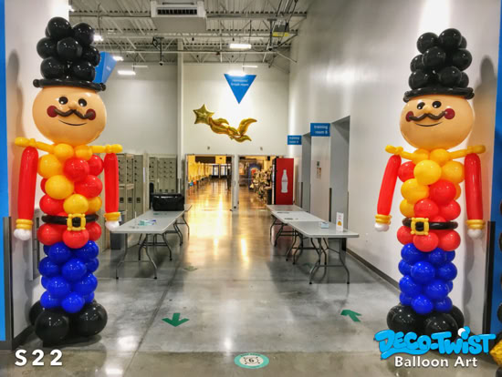 Two large balloon sculptures of toy soldiers stand on either side of a hallway entrance. Towering 10' high – each figure has a black hat, a smiling face with a mustache, red arms, yellow and red chest, blue legs, and black boots.