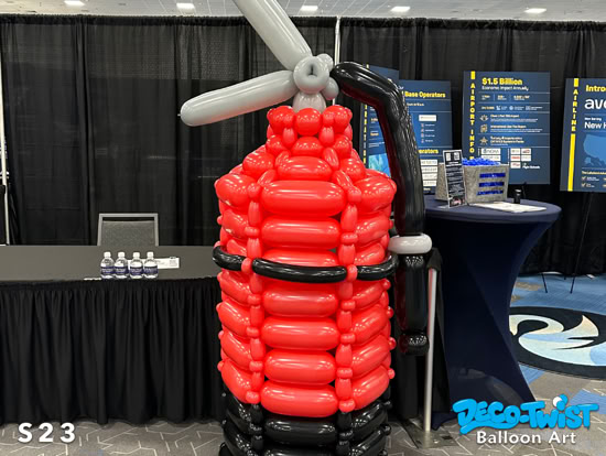 A large balloon sculpture shaped like a fire extinguisher, made of red, black, and gray balloons with a balloon nozzle and handle on top.