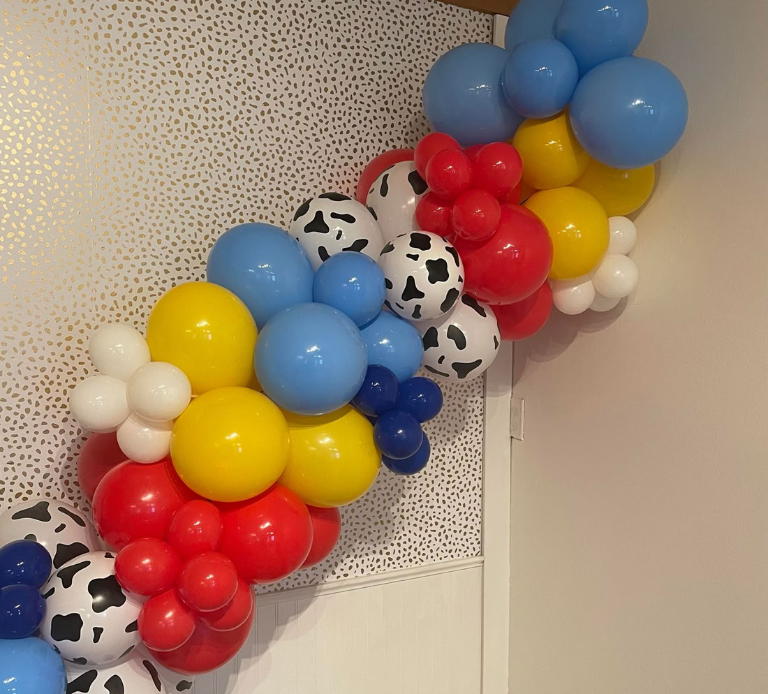 This image features a vibrant balloon garland mounted on a wall. The garland is made up of shiny balloons in primary colors: red, yellow, blue, and white, along with a few balloons that have a black-and-white cow print pattern. The backdrop is a light wall with a subtle gold dotted pattern. The overall design gives off a cheerful, fun vibe, likely inspired by a Toy Story theme, perfect for a birthday or children’s party.