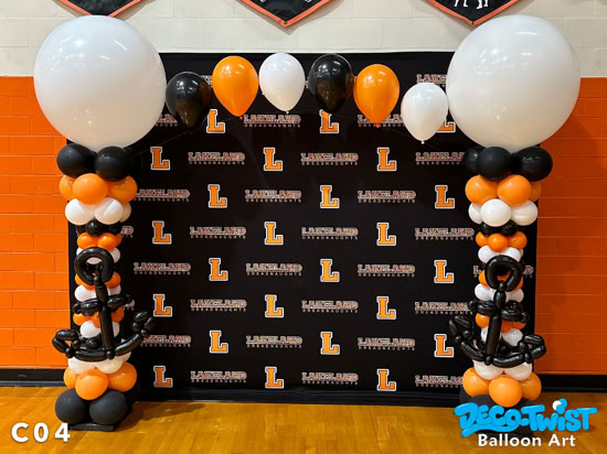 Two balloon columns frame a Lakeland Dreadnaughts backdrop. The columns are decorated in black, orange, and white balloons with large white balloons on top. Midway down, each column features a balloon anchor design, and a string of orange, black, and white balloons connects the tops across the backdrop.