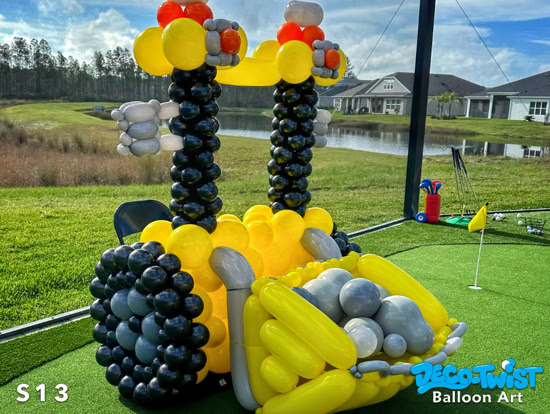 A large balloon sculpture depicts a yellow construction front loader. It has black balloon wheels, a cab structure, and a gray and yellow balloon scoop filled with round balloon “rocks.” A perfect photo opportunity for construction themed a party or event.