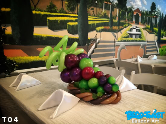 This image shows a creative balloon centerpiece shaped like a bunch of grapes. The arrangement includes clusters of round balloons in shades of green, purple, and red to resemble grapes, with long green balloons forming vine-like stems at the top. At the base, brown twisted balloons create a basket effect. The centerpiece is displayed on a decorated dining table with neatly folded white napkins, giving it an elegant and playful look.