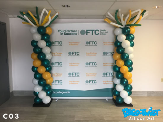 Two tall balloon columns stand side by side in front of a Florida Technical College banner. The columns are spiraled with green, yellow, and white balloons and topped with sprays of long, thin balloons bursting upward like fireworks, creating a celebratory effect.