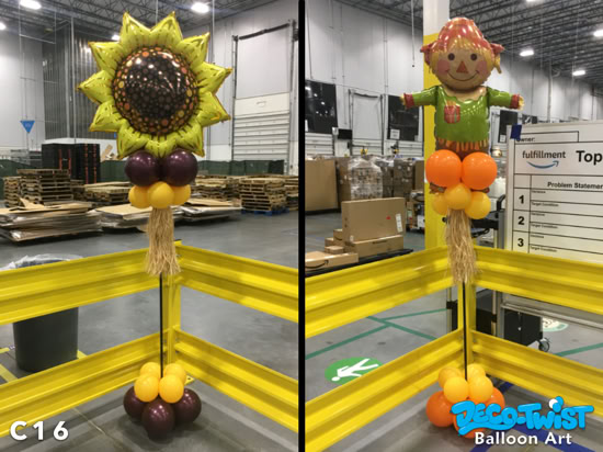 This image shows two balloon columns with fall-themed toppers. On the left, the column is topped with a large sunflower balloon, with clusters of brown and yellow balloons beneath it and raffia accents resembling straw. On the right, the column features a scarecrow balloon topper, supported by orange and yellow balloon clusters with matching raffia details. Both columns are designed to resemble harvest or autumn decorations, placed in an warehouse setting.