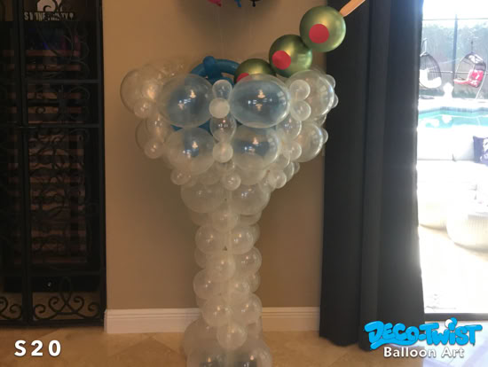 A balloon sculpture shaped like a martini glass is shown, made of clear balloons with blue accents. It’s topped with balloon olives on a stick for garnish.