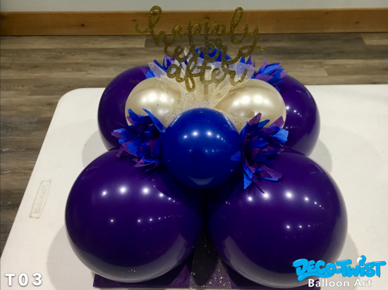 This image shows a custom desinged balloon centerpiece featuring a large purple balloons forming the base, accented with smaller pearl-white and blue balloons. Blue tissue paper decorations are tucked in for added flair. At the top, a glittery gold sign reads “Happily Ever After,” making it a perfect choice for the head table at wedding reception.