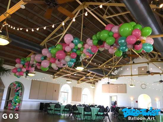 A large balloon garland made of pink, green, teal, and white balloons is suspended from the ceiling of the Magnolia Building in Lakeland, Florida adding festive color above round tables set for guests.