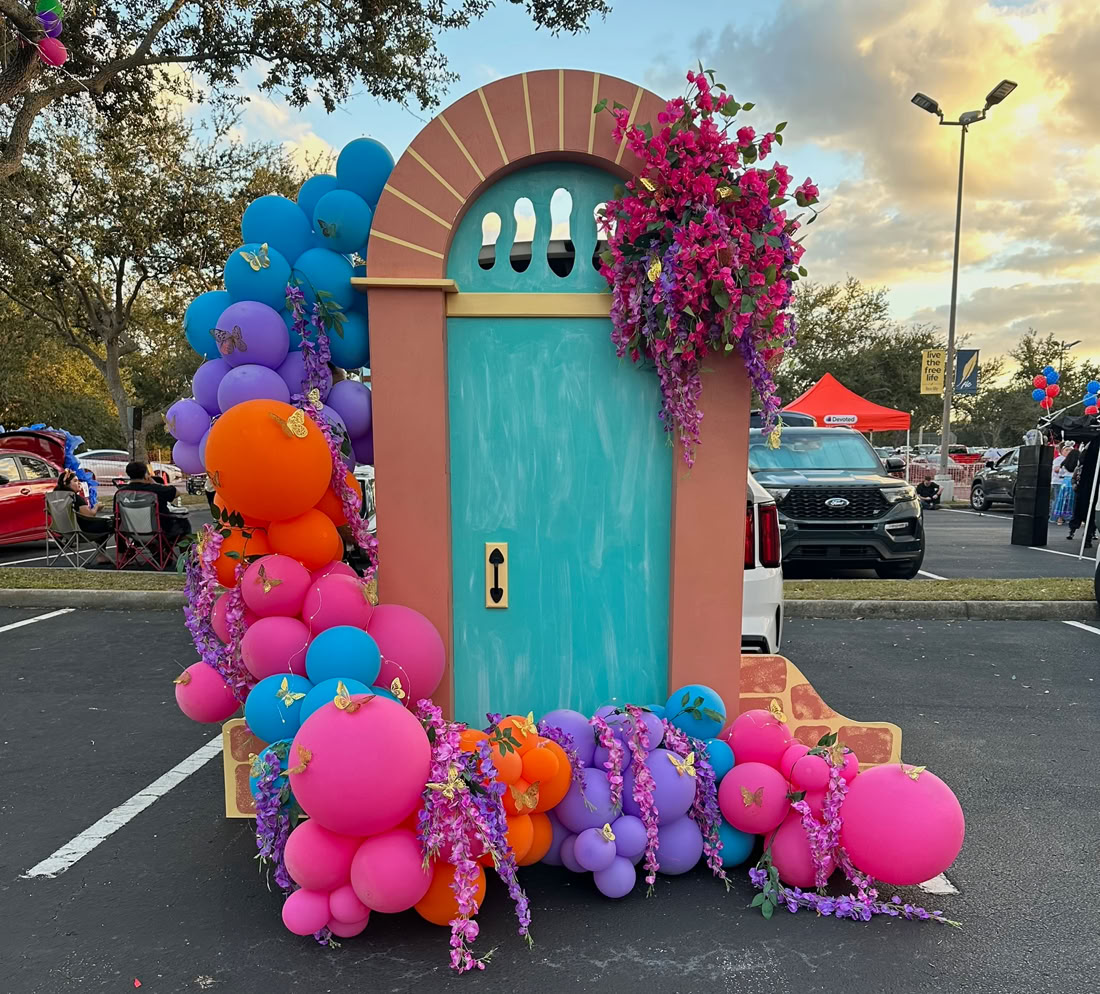 This image shows a whimsical display set up in a parking lot, featuring a large decorative door that is turquoise with a rounded top and coral-orange frame. The door is accented with vibrant pink and purple faux flowers spilling over the top and around the base. Colorful balloon clusters—mainly pink, orange, purple, and blue—surround the door on both sides, creating a festive and playful look. The scene suggests a themed event or celebration, with people and cars visible in the background under a partly cloudy sky.