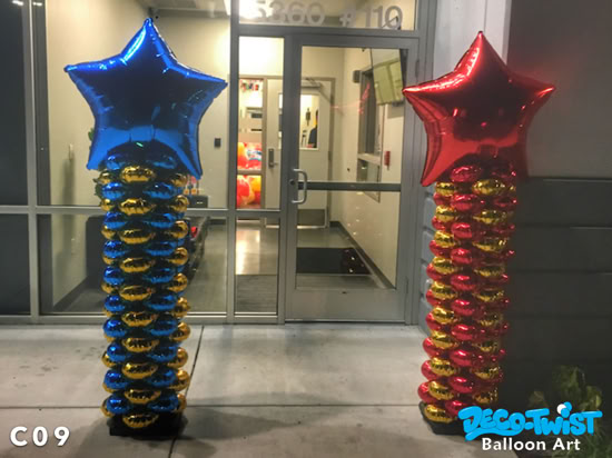 This image shows two tall balloon columns placed outside a building entrance. Each column is made of stacked shiny foil balloons in alternating colors, with one column featuring gold and blue balloons topped with a large metallic blue star, and the other featuring gold and red balloons topped with a metallic red star. The display is brightly reflective and eye-catching, designed as festive balloon art for an event entrance.