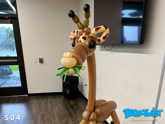 A tall balloon sculpture of a giraffe stands indoors. The giraffe is made of brown balloons with patterned ears, big eyes, and green balloons in its mouth to look like it’s eating leaves.