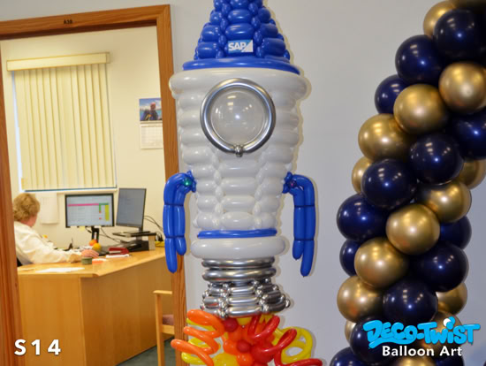 A balloon sculpture of a rocket stands indoors, made of white, blue, and silver balloons with orange and yellow balloons at the bottom to look like flames. The rocket has a round window in the center.