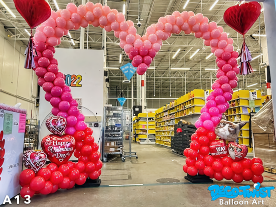 A large heart-shaped balloon arch made of red, dark pink, and light pink balloons creates a festive display. The base is decorated with red balloons and Valentine’s Day-themed heart balloons, including ones that say “Happy Valentine’s Day.”