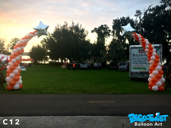 This image shows two tapered balloon columns outdoors, each spiraling upward in orange and white balloons. At the top of both columns are large silver star-shaped balloons, giving the display a festive and celebratory look. The columns perfectly frame the entrance to Anchor Park in Lakeland, Florida.