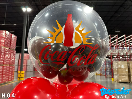 This image shows a clear helium balloon with black balloons inside and a red Coca-Cola Beverges of Florida logo in custom vinyl on the outside. The balloon is part of a bouquet, with red balloons visible beneath it. It is displayed indoors in what looks like a warehouse or event space.