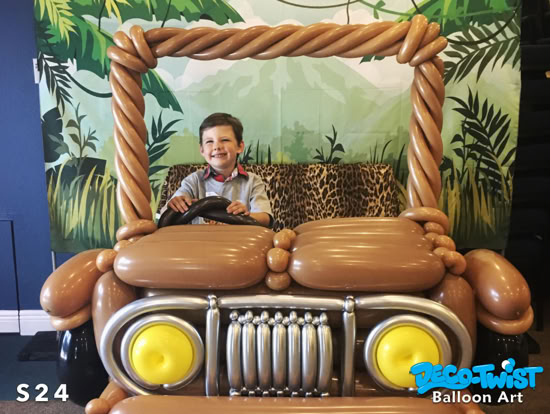 A young boy is sitting inside a balloon photo oportunity shaped like a brown Jeep. The Jeep has balloon headlights, a front grill, and a steering wheel, with a jungle-themed backdrop behind him.