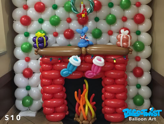 A festive balloon sculpture depicts a Christmas fireplace. Red balloons form the brick mantel, with stockings hanging in front, a balloon fire inside, and wrapped presents, a wreath, and a balloon dog on top. White, red, and green balloons create a holiday-themed backdrop behind the fireplace.