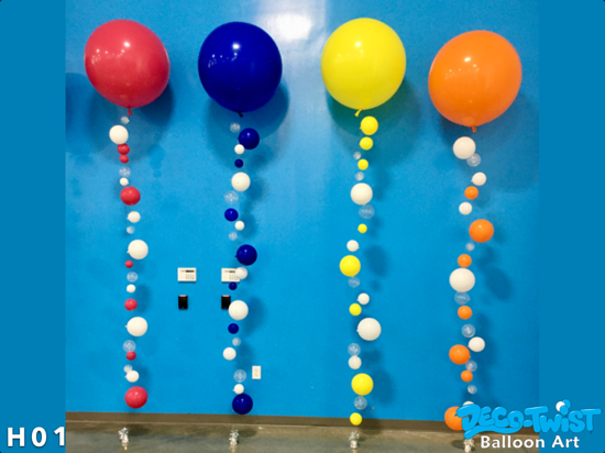 This image shows four large helium balloons in red, blue, yellow, and orange, each with a long string of smaller balloons attached beneath them. The balloon strands are made up of different-sized balloons in matching colors, alternating with clear balloons, giving them a bubbly, floating appearance. The setup is against a bright blue wall, creating a playful and colorful display.