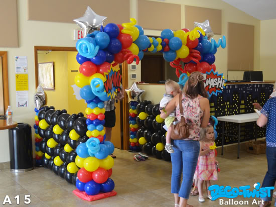A colorful superhero-themed balloon arch frames a doorway at an indoor event. The arch is built from bright red, yellow, blue, and black balloons, with fun curly balloon accents, silver star balloons, and comic-book style signs that say “WHAM!” and “SMASH!” Nearby, children and adults walk under or around the arch.