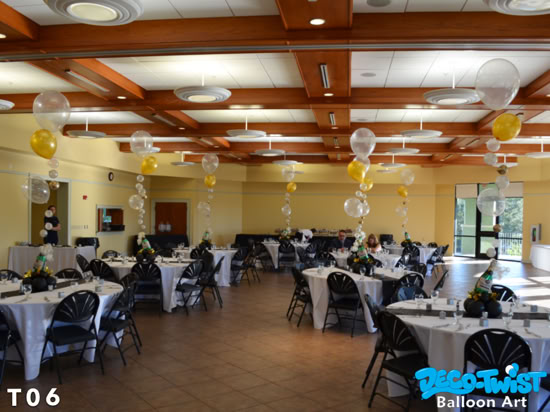 This image shows a decorated banquet hall with round tables set for an event. Each table has black chairs and a centerpiece featuring a champagne bottle balloon surrounded by smaller balloons and flowers. Along the center of the room, tall balloon arrangements rise from the floor, featuring large gold and clear balloons with smaller balloons stacked below them in a vertical column. The overall color scheme is gold, black, and white, giving the space an elegant and celebratory atmosphere.