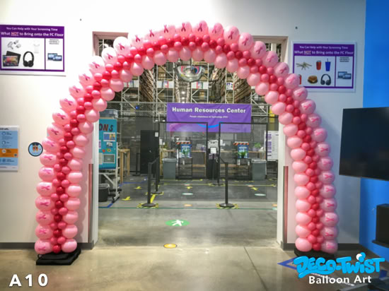 A large pink balloon arch stands in front of a workplace entrance. The arch is made of light and dark pink balloons, with some featuring printed pink ribbons, symbolizing breast cancer awareness.