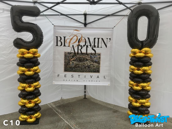 Two tall latex-free balloon columns stand side by side, made of alternating shiny gold and black balloons. At the top of the columns are large black foil number balloons forming the number "50." Behind them is a banner reading Bloomin’ Arts Festival Bartow, Florida, celebrating the festival’s 50th anniversary.