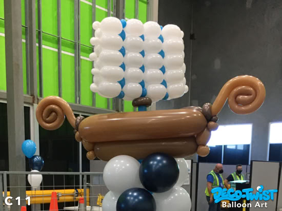 This is a balloon sculpture designed to look like a viking ship. The base is made of stacked blue and white balloons, supporting a brown balloon structure shaped like a boat with curled ends. Above it, white and blue balloons form a square sail, completing the nautical theme.