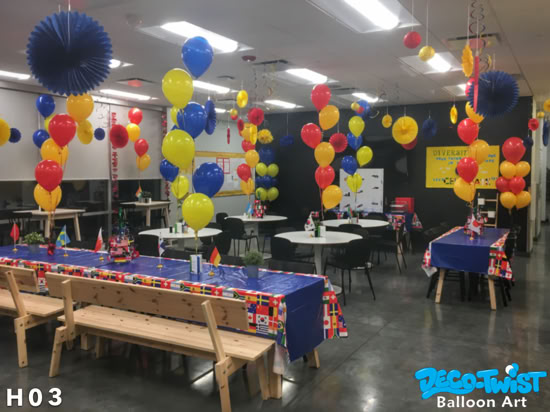 This image shows a lively event space decorated with red, yellow, and blue helium balloon bouquets placed at tables throughout the room. Each bouquet has three balloons tied together and floating above the tables, which are covered with blue tablecloths and flag-themed accents. Hanging paper decorations in matching colors add to the festive atmosphere, creating a cheerful and vibrant celebration.
