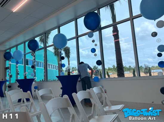 This image shows a row of tall helium balloon arrangements set up indoors for an event. Each bouquet features a large balloon on top in shades of blue, silver, and white, with strings of smaller balloons cascading down the ribbons. The decorations line tall windows that let in natural light, while white folding chairs and blue cocktail tables are arranged in the room for guests. The setup gives a clean, modern, and celebratory look.
