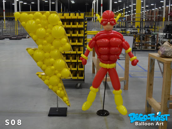 A balloon sculpture of the superhero Flash stands next to a large yellow lightning bolt made of balloons. The figure is red with a muscular chest design, yellow boots, and a mask.