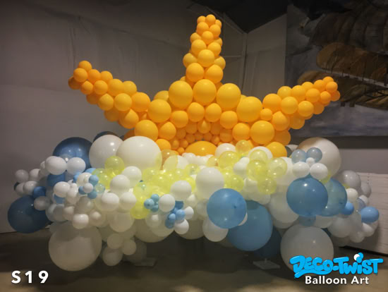 A large balloon sculpture shows a bright yellow sun rising above fluffy clouds made of white, blue, and light yellow balloons, creating a cheerful sky scene.